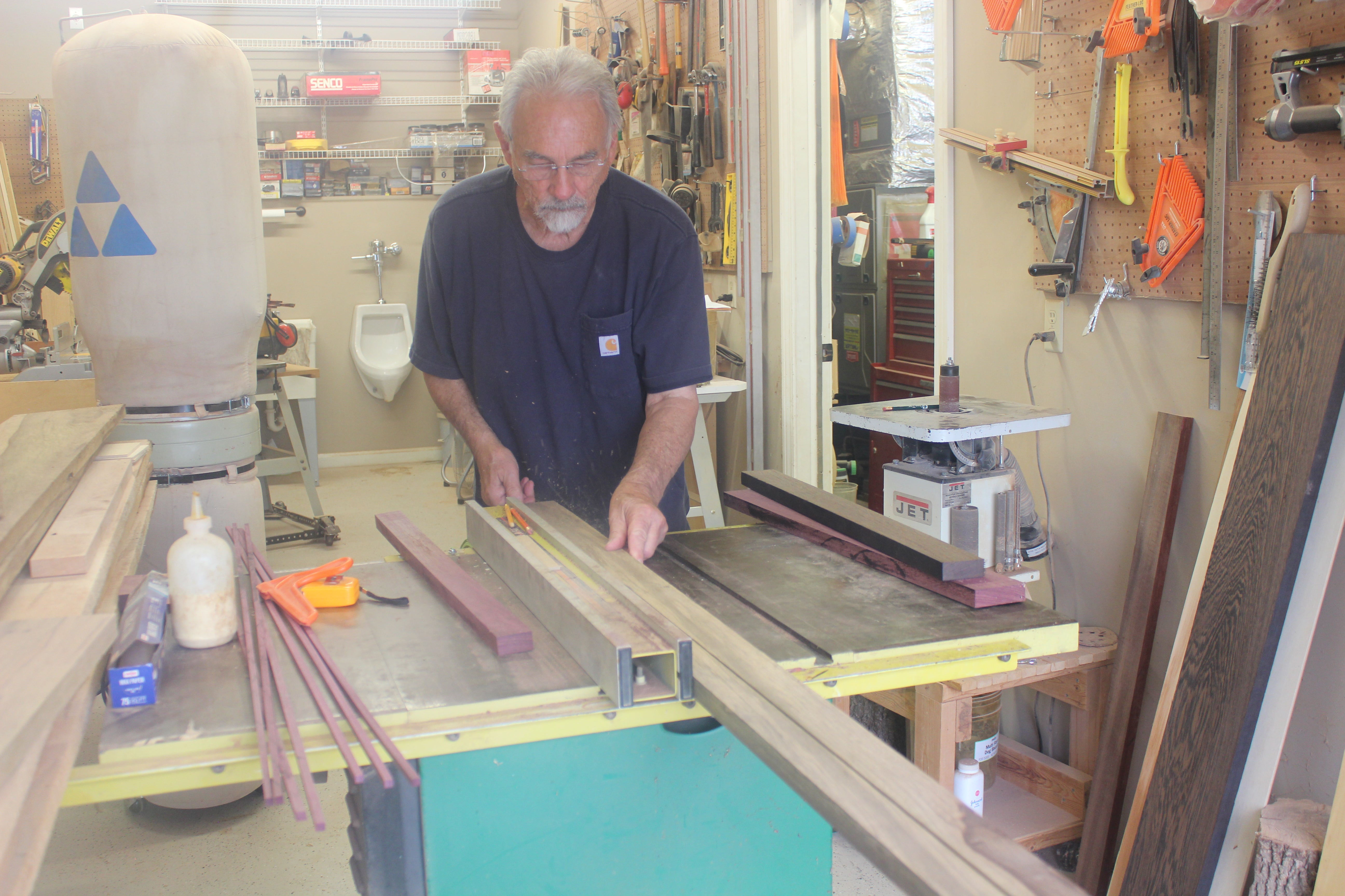 Man operating a table saw in a workshop with various tools and equipment around.