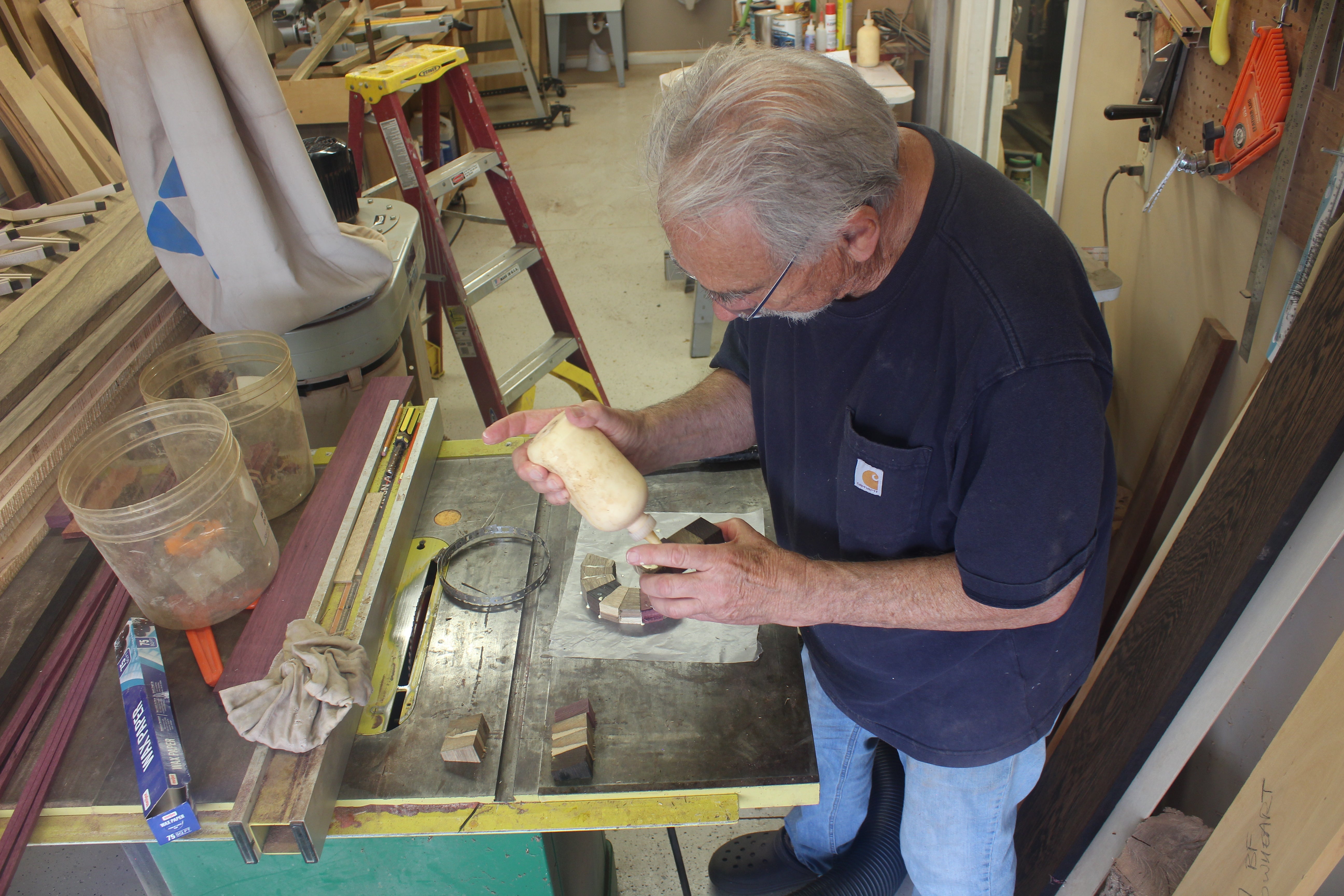 Man working on a project in a workshop with tools and materials. Huntertown, Indiana woodworking shop with Thom looking at a glue bottle pieces.