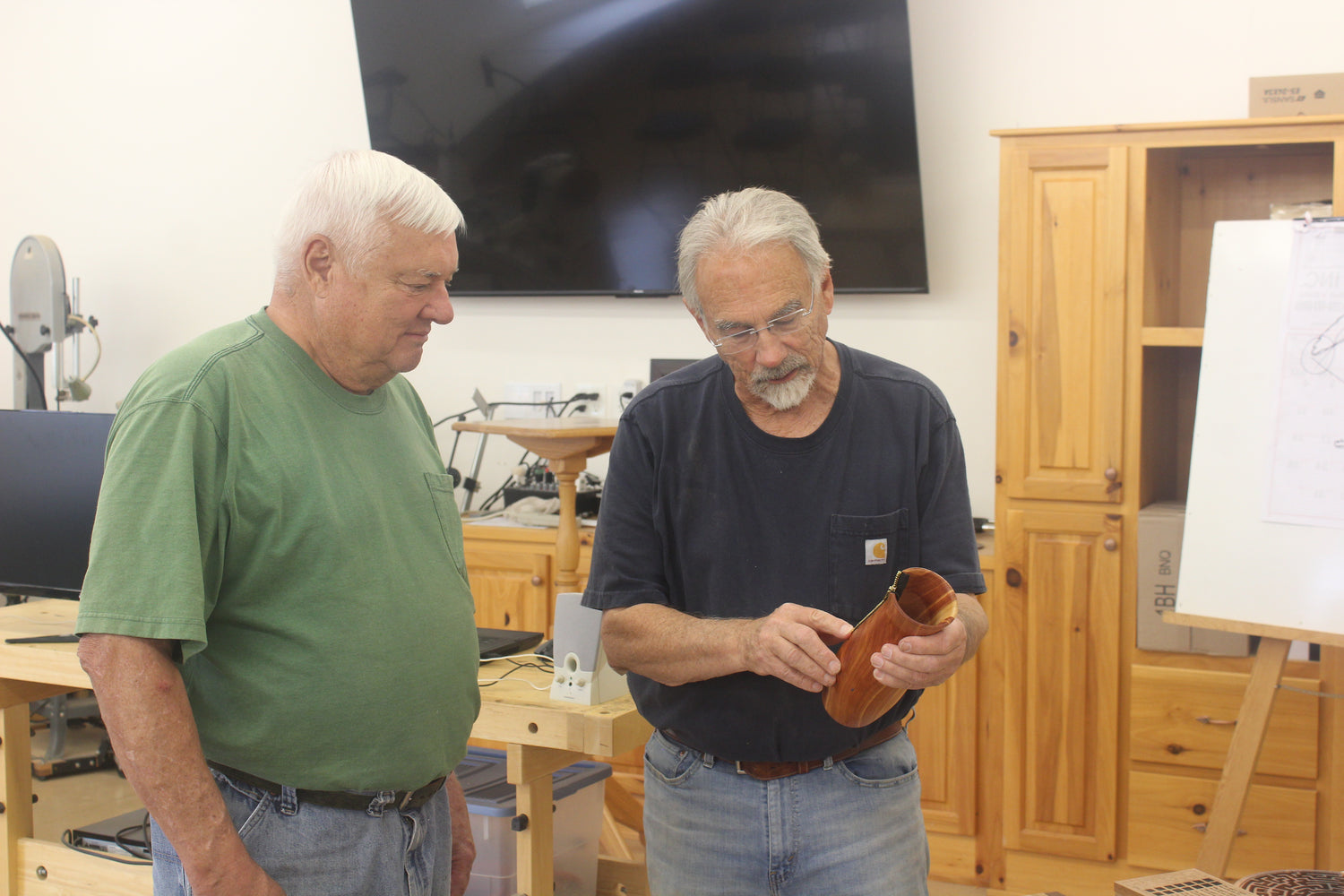 Two men in a workshop setting, one holding a wooden object. Thom Pabst explaining the information regarding a unique wooden piece. A joyful friend is looking at it.