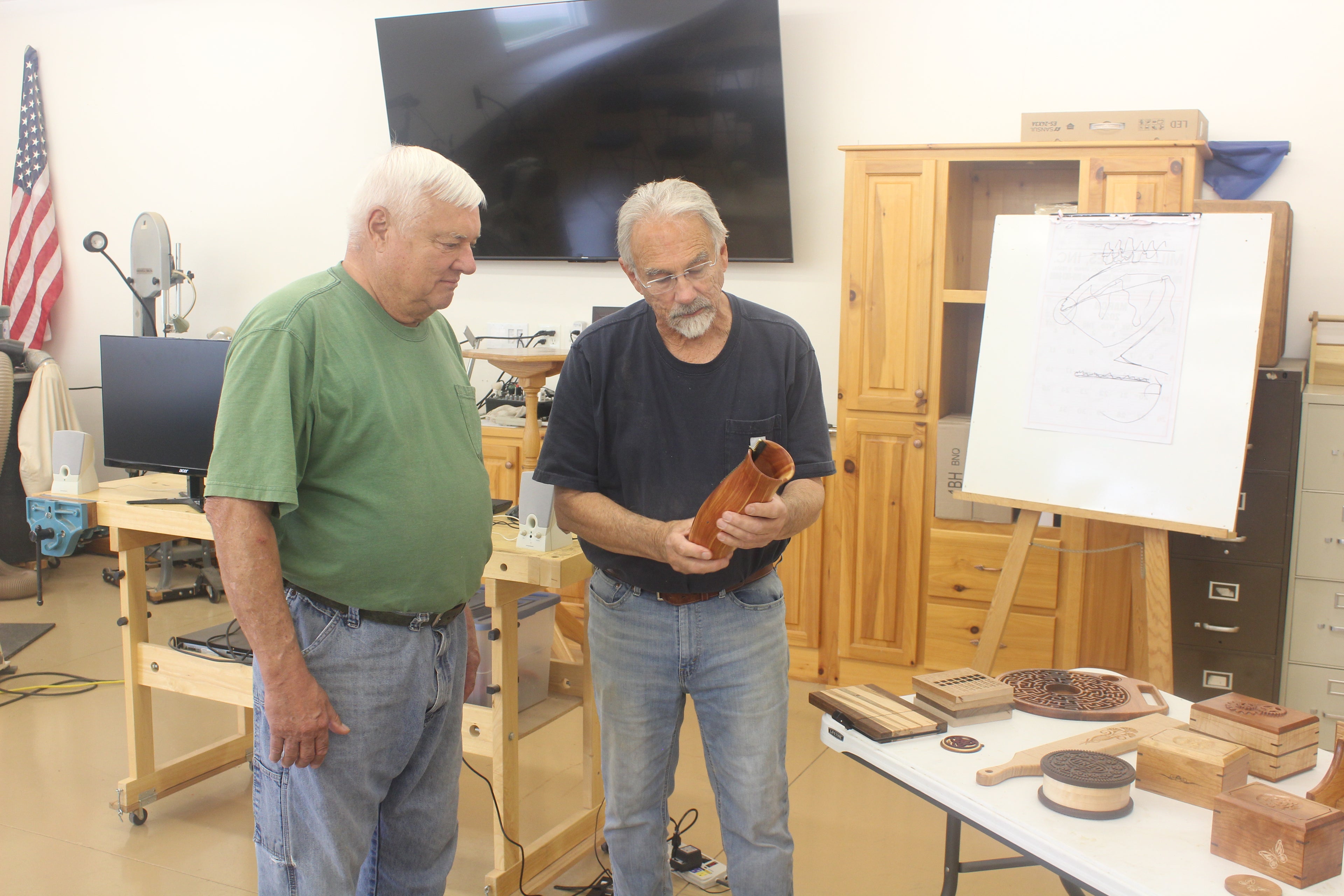 Two men in a workshop setting, one holding a wooden object, with tools and equipment around. It is taking place in a wood working shop in Huntertown, Indiana, United States.