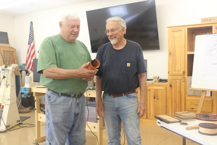 Two men standing in a workshop with tools and equipment around. Huntertown, Indiana woodworking shop with member looking at wooden pieces.