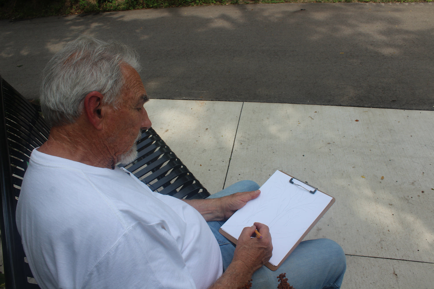 Man sitting on a bench outdoors, drawing on a clipboard.